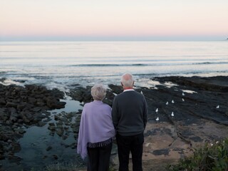 An older couple standing together on a rocky shoreline, gazing at the calm ocean under a pastel sky at dawn or dusk, surrounded by seagulls in a serene and peaceful setting