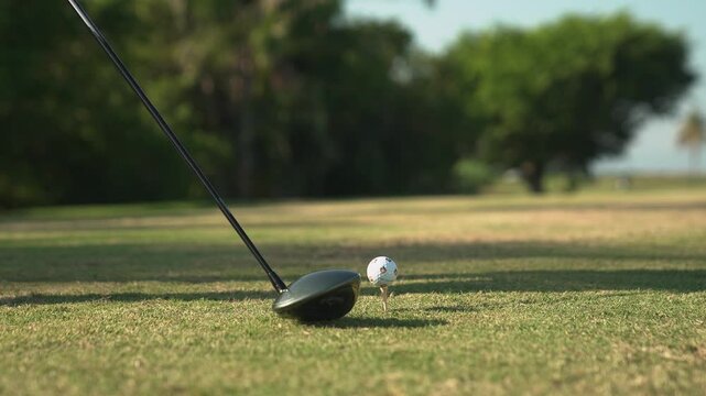 A professional golfer takes a powerful swing with a driver, hitting a golf ball off the tee on a lush green golf course. The video captures the moment of impact in clear detail, set against a scenic b
