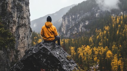 Man sitting on a rock overlooking a valley. He is wearing a yellow jacket and black beanie. The valley is filled with trees and fog.