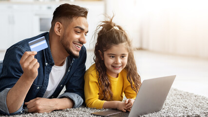 Portrait of happy arab dad and cute little daughter with laptop and credit card making online...