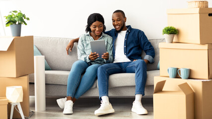 Happy Black Couple Using Digital Tablet Together While Sitting On Couch After Moving To New Home, Cheerful African American Spouses Shopping Online On Tab Computer Or Browsing Internet, Copy Space