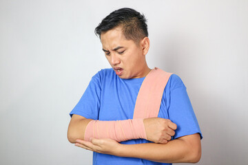 Handsome young asian man with broken arm in soft splint suffering a sore arm isolated on grey background, accident insurance concept.