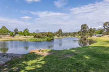 Fototapeta premium Quart Pot Creek with sunny and shady grass banks in a park scene, flows through the centre of the town of Stanthorpe in Queensland, Australia, under a blue sky with clouds.
