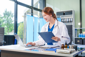 A young Asian female doctor, confidently dressed in a white uniform, sits at a computer desk in a...