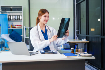A young Asian female doctor, confidently dressed in a white uniform, sits at a computer desk in a...