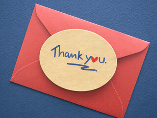 Top view, flat lay. Positive emotion concept. Thank you card. Close-up "thank you" message is handwritten on a brown oval paper on red envelope over dark blue background..