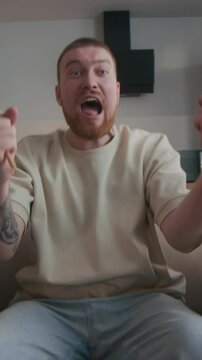 Vertical medium slowmo of focused Caucasian man sitting on couch in front of TV watching sports, shouting and cheering emotionally for winning team and spinning wooden ratchet noise maker grogger