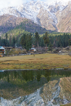 Snowy Nanga Parbat reflects perfectly in a calm lake at Fairy Meadows, Pakistan. Surrounded by pine forests, wooden cabins, and a serene meadow.