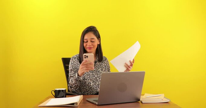 Smiling Asian working woman using smartphone hold documents read sms on social media looking happy at workplace, isolated over yellow background. woman searching e-commerce offers on cell phone.