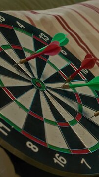 Vertical closeup of unrecognizable players throwing darts at target on board while playing in cozy apartment with striped cusion on couch