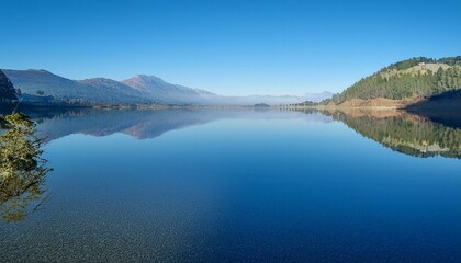 A tranquil lake reflecting a clear blue sky and distant mountains, captured in soft light.
