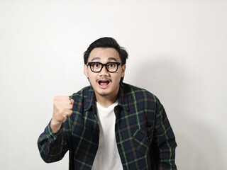A young Asian man in a plaid shirt pumps his fists in excitement and smiles broadly against a solid white background