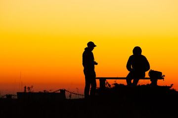 Two people silhouetted against a vibrant orange sunset quietly on a bench at a scenic overlook. The silhouettes contrast beautifully with the colorful sky, creating a serene atmosphere.