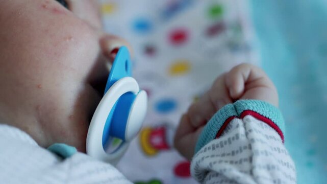 A small caucasian child sucks on a bare pacifier, the focus shifts from the pacifier to a small hand with a fist, close-up
