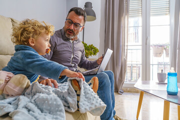 Father managing work and family life, using laptop while attending his toddler son in cozy living room