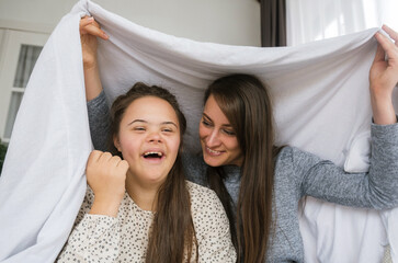 A young woman with Down syndrome and her mother share a joyful, playful moment under a white sheet....