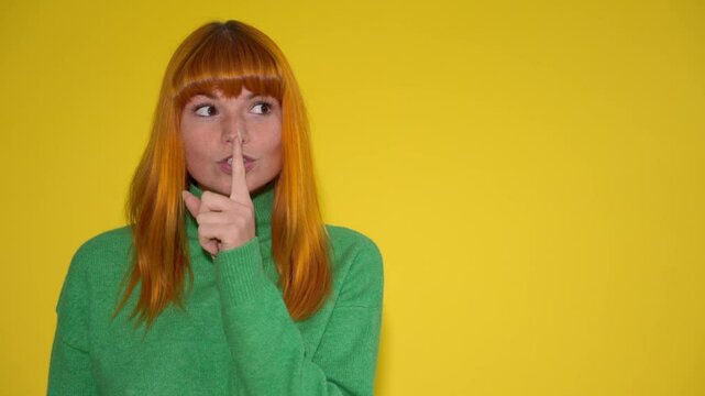 Woman with orange hair and green sweater holding finger to lips in shush gesture against a bright yellow background