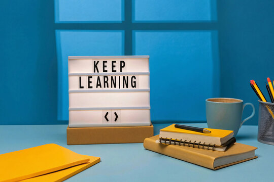 A minimalist desk features a lightbox reading KEEP LEARNING, notebooks, pens, and a coffee cup, set against a window-lit blue backdrop