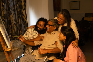 young indian family of four enjoying with senior disable father sitting on wheel chair painting on canvas with a brush and palette at home.