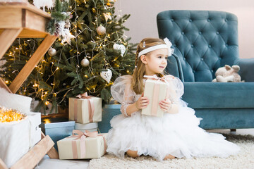 Little girl with blond hair in white dress sitting near Christmas tree holding a gift and smiling.
