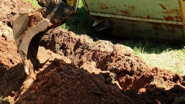 Mini excavator digs a narrow trench in soil for laying communications close-up. Mini bucket of crawler machine. Earthworks with heavy special equipment. Digging machinery works at construction site.