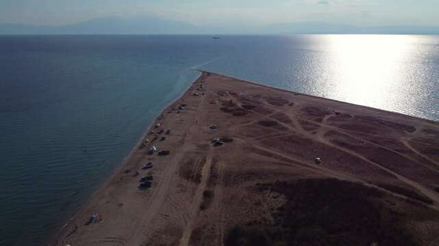 Forward drone shot captures a tranquil beach peninsula, with calm blue waters and shimmering sunlight reflected on the vast sea. Sandy beach trails, scattered campers, and untouched coastal beauty.