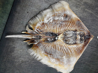 A dried ray fish, flat and wide, with its fins spread out, shows a smooth texture and light brown color. © Magdalena