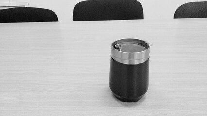 a black and silver insulated travel mug placed on a light-colored wooden table. The mug has a black body with a silver top, likely made of stainless steel. In the background