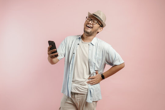 Happy young Asian man wearing hat and blue shirt is laughing while holding his smartphone.