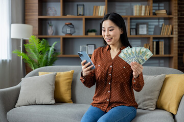 Asian woman sitting on sofa with smartphone in hand, showing excitement while holding cash. Bright, modern home interior, depicting success, financial achievement, happiness and positivity.