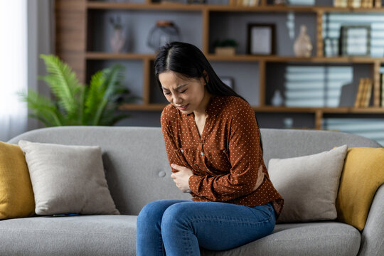 Asian woman on sofa holds stomach with discomfort. Suffering from stomachache or digestive issues. Casual indoor setting at home conveys pain and discomfort.