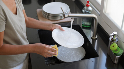 Woman washing and rinsing dishes by hand under a stream of water in the kitchen sink. Washing...