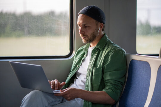 Focused businessman type on laptop, wearing earphones while traveling by train to work. Mobile office setup, digital nomad lifestyle, remote workspace, tech savvy freelancer, commute with productivity