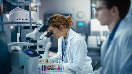 Medical Research and Development Laboratory: Beautiful Hispanic Female Scientist Working on a Computer and Looking at a Sample in a Petri Dish Under a Microscope. Advanced Biotechnology Lab