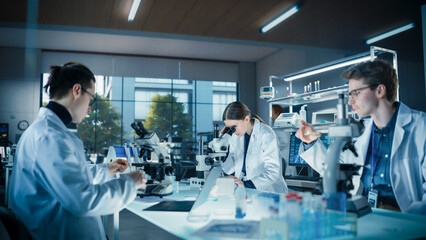 Young Team of Medical Researchers Sitting Behind a Table, Using Microscopes for Medical Pharmaceutical Research and Treatment Development. Specialists Working in a Modern Laboratory