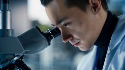 Portrait of a Handsome Chemistry Student Working on a Research Project in University. Medical Research Scientist Looking at Biological Samples Under a Microscope in an Applied Science Laboratory