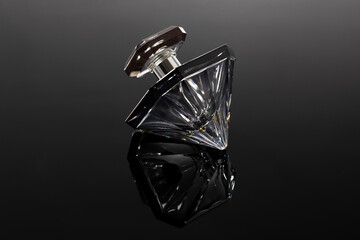 An elegant diamond-shaped glass perfume bottle is tilted on a sleek black reflective surface. The...