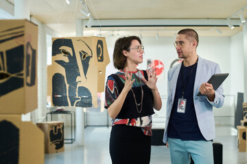 Young female artist talking to gallery worker with tablet while preparing for opening exhibition of...