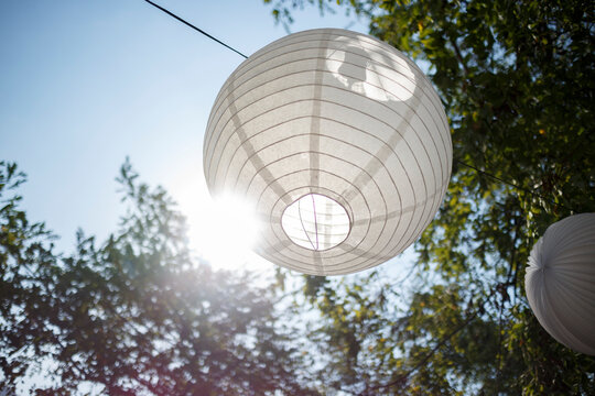 A large, light-gray paper lantern hangs amidst lush green trees against a partly cloudy sky.  Sunlight shines through the lantern.