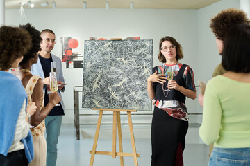 Young owner of art gallery holding glass of champagne and communicating to visitors of exhibition...