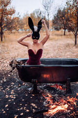 A girl in a burgundy bodysuit sits in a warm bath with water heated over a fire