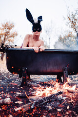 A girl in a burgundy bodysuit sits in a warm bath with water heated over a fire