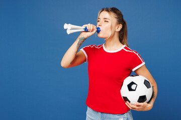 Side profile view young woman fan wear basic red t-shirt cheer up support football sport team hold in hand soccer ball watch tv live stream blowing pipe look aside isolated on plain blue background.