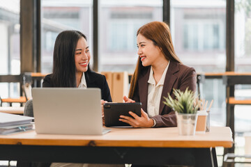 Two businesswomen in formal attire discussing work while using tablet at modern office desk with laptop and potted plant, smiling and engaging in professional conversation