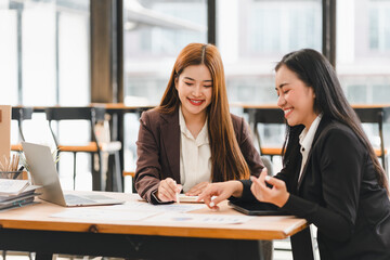 Two businesswomen in formal attire discussing documents at wooden desk in modern office with large windows, smiling and collaborating on project