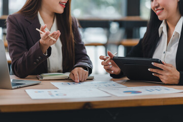 Two businesswomen discussing financial reports at wooden desk with laptop and tablet, showcasing teamwork and collaboration in modern office