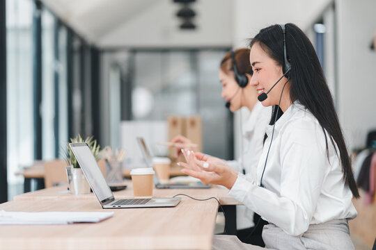 Two female customer service representatives wearing headsets work at desks with laptops, engaging in professional communication in modern office space