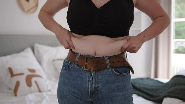 Woman concerned about her belly fat, squeezing them with hands wearing jeans and belt in a casual bedroom setting
