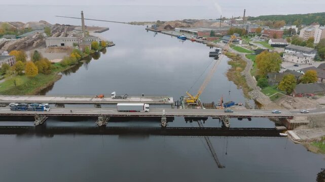 Aerial establishing shot of traffic crossing bridge connection over Salaca river (Salacgriva city) near Estonia and Latvian border, construction site with cranes and cargo transport