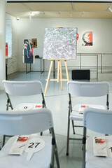 Part of spacious gallery hall with abstract painting and rows of chairs for auctioneers with brochures and paddles with numbers